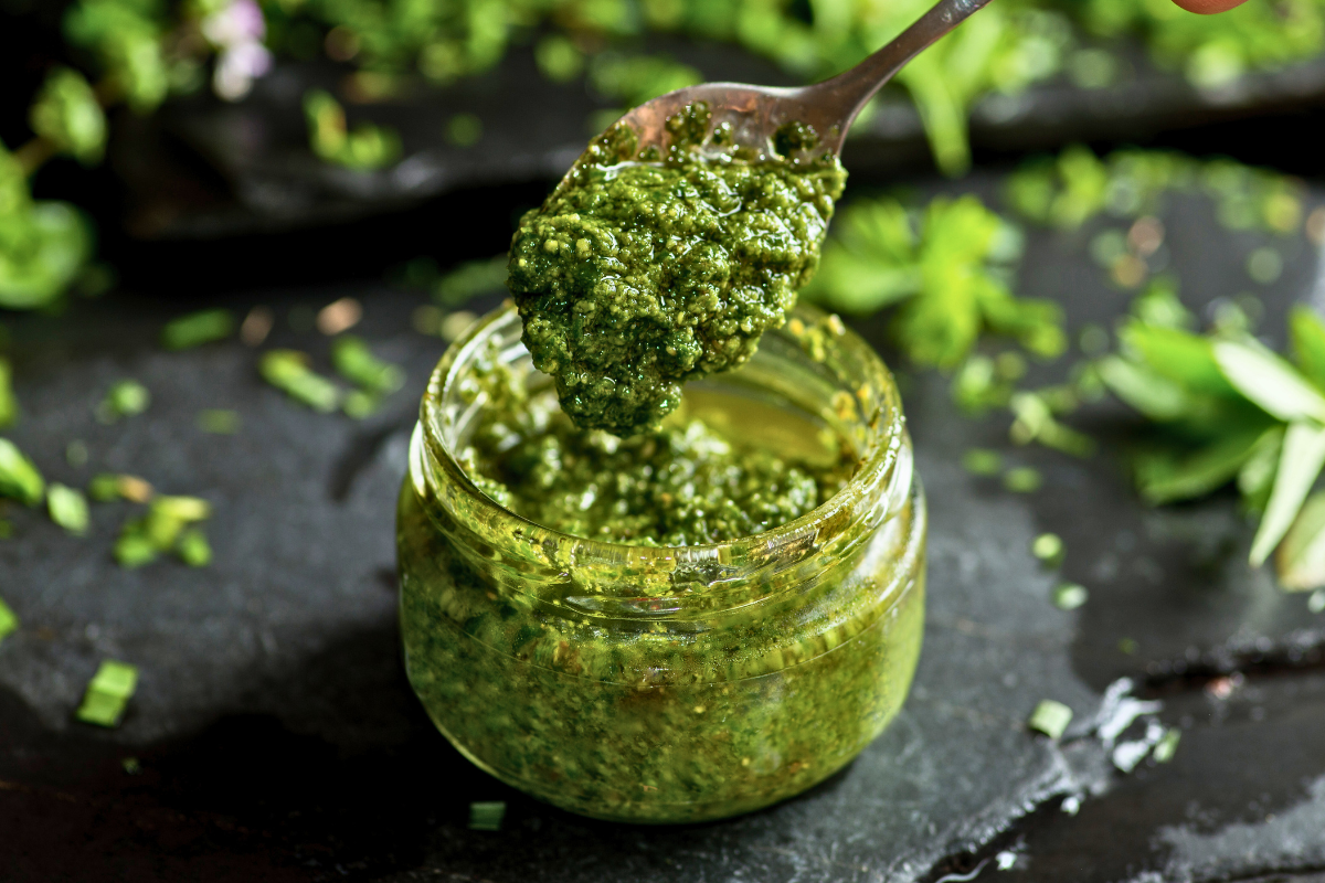 Jar of green pesto with a spoonful above on a dark surface